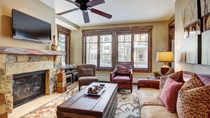 A living room with a fireplace, a ceiling fan, a television, and a large window with a view of buildings. - Centrally located ski chalet with shared hot tub and pool - right in downtown (Breckenridge)