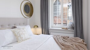 Ein ordentlich bezogenes Bett mit weißen Leinen und einer beige farbenen Tagesdecke, ein runder Spiegel an der Wand und ein Fenster mit grauen Vorhängen.