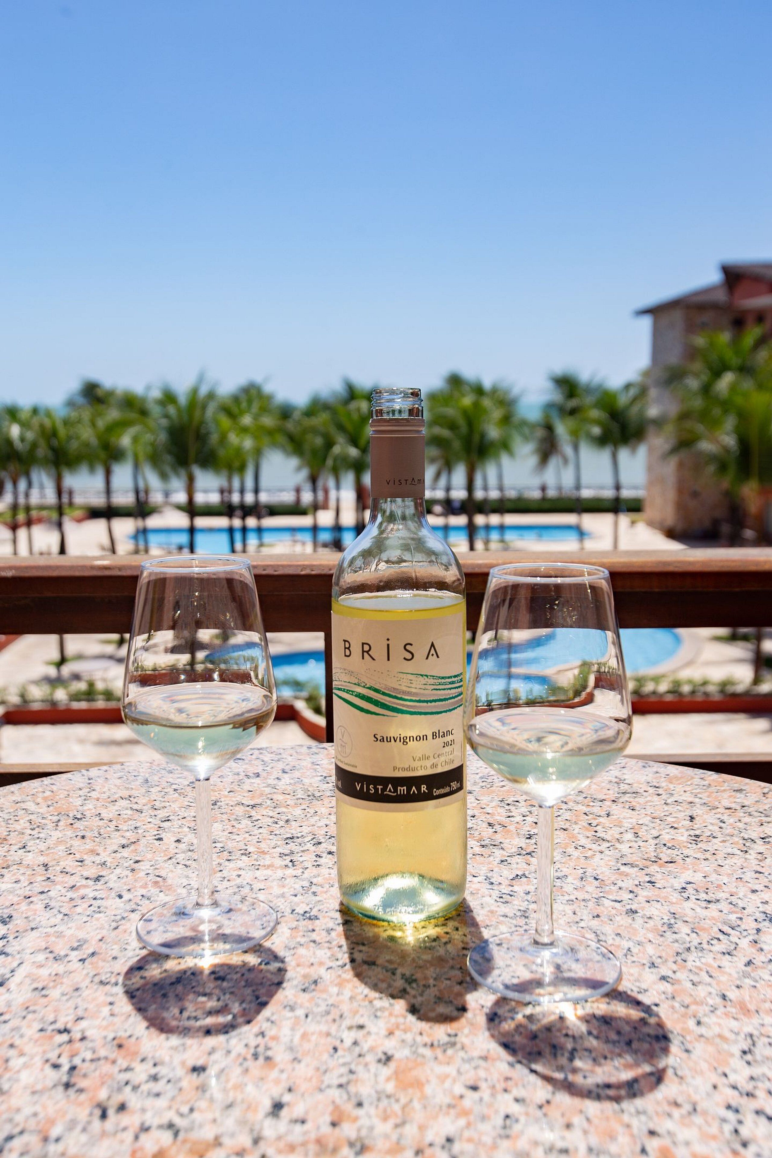 Een fles Brisa Sauvignon Blanc en twee glazen op een tafel, met een zwembad en palmbomen op de achtergrond.