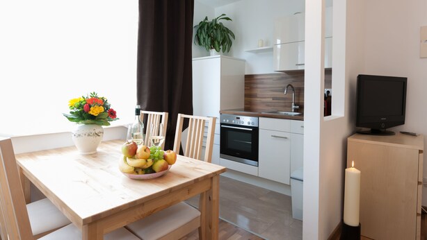 A modern kitchen with white cabinets, a wooden dining table with a fruit bowl, and a TV on a cabinet. - Rafael Kaiser - Apartment Cityflow (Graz)