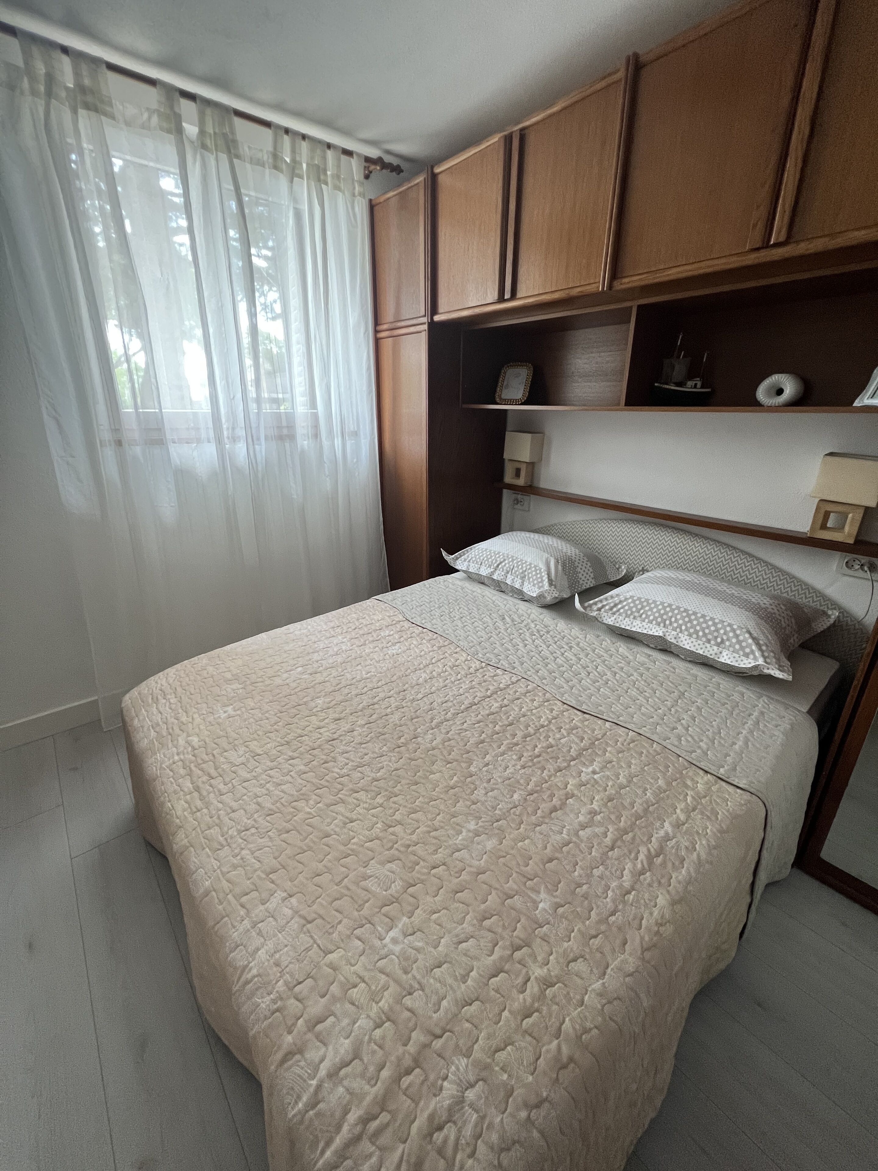 A bedroom with a bed, wooden cabinets, a nightstand, and a window with sheer curtains.