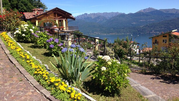 Un jardín en la ladera con diversas flores, un edificio de madera con balcón y vistas a montañas y un lago.