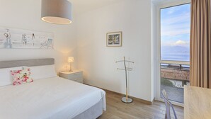 Habitación de hotel con cama, mesita de noche, lámpara, perchero, ventana con vistas y una obra de arte en la pared.