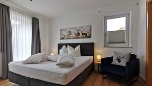 Ein Schlafzimmer mit einem großen Bett, einem Nachttisch mit Lampe, einem dunklen Sofa mit Kissen in Hirschoptik und einem Fenster mit Jalousien.