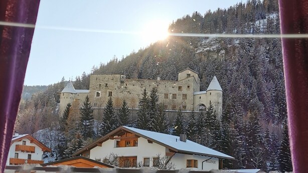 Blick auf ein schlösserartiges Gebäude, umgeben von schneebedeckten Bäumen, mit blauem Himmel und Sonnenschein.
