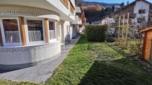 Ein Wohngebäude mit einem gebogenen Balkon, einer kleinen Gartenanlage mit Hecke und Blick auf eine hügelige Landschaft im Hintergrund.