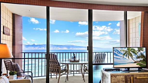 Ein Balkon mit Meerblick, ausgestattet mit Stühlen und einem Tisch, sowie ein Fernseher auf einer Holzkommode.
