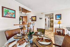 A dining area with round tables set for four, surrounded by wicker chairs. There are framed artworks on the walls, a window with a view, and a door leading to another room.