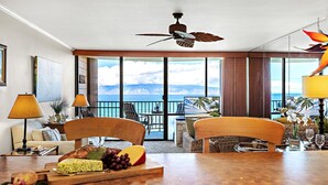 Ein Hotelzimmer mit einem Holztisch, zwei Stühlen, einem Deckenventilator, einem großen Fenster mit Meerblick und einem Balkon mit Sitzgelegenheit.