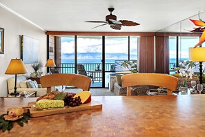 A hotel room with a wooden dining table, two chairs, a ceiling fan, a large window with a view of the ocean, and a balcony with outdoor seating.