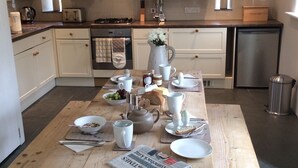 A modern kitchen with white cabinets, a wooden dining table set with breakfast items, and a newspaper on the table. - Peaceful Rural Retreat, Ideal Base To Explore Cornwall (Wadebridge)