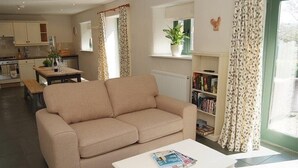 A living room with a beige sofa, a coffee table with magazines, a bookshelf, and a window with curtains. - Peaceful Rural Retreat, Ideal Base To Explore Cornwall (Wadebridge)