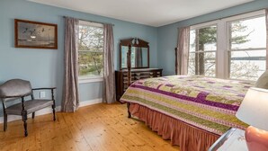 Une chambre avec un plancher en bois, un lit recouvert d’une courtepointe aux couleurs vives, une chaise, une commode et une fenêtre donnant sur des arbres et l’eau.