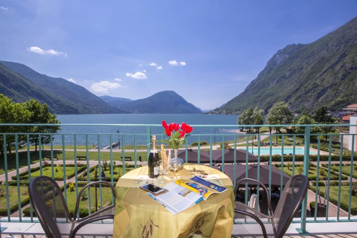 A table set with a bottle, glasses, and a menu on a balcony overlooking a lake with mountains in the background.