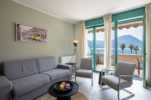 A modern living room with a sofa, chairs, a coffee table with drinks, a floor lamp, and a painting of a tram. - Palazzo Apartments - Porto Letizia (Porlezza)