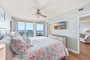Un dormitorio con cama, ventilador de techo, ventana grande con vista al océano, un cuadro en la pared y mesita de noche con lámpara.