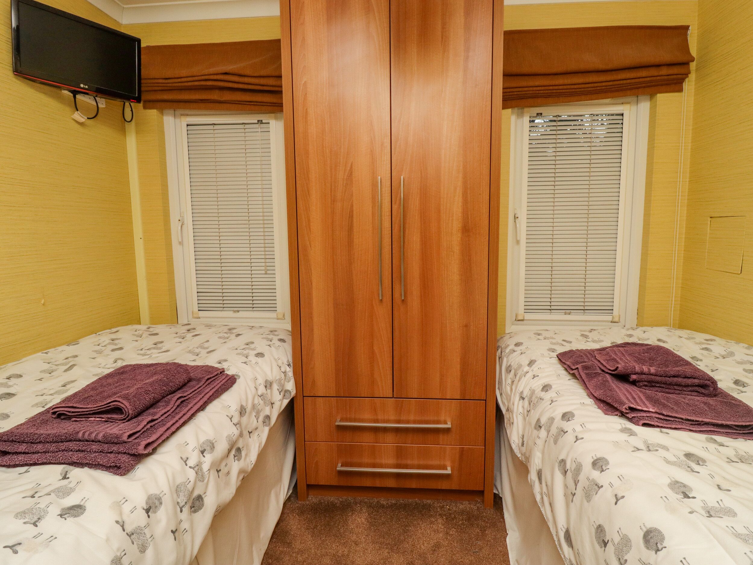 A bedroom with two beds, a wooden wardrobe, and a TV mounted on the wall.