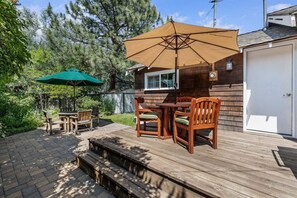 A wooden deck with a table and chairs, a green umbrella, and a wooden building in the background. - Bluebird Day - Desert Moon, Cozy 2BR in Bend's River West, Walkable to restaurants! (Bend)