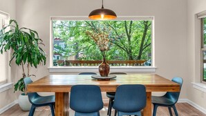 A dining area with a wooden table, four blue chairs, a hanging pendant light, and a large window with a view of trees and a fence. - Bluebird Day - Hartford Haven, a spacious 3BR home with modern amenities. (Bend)