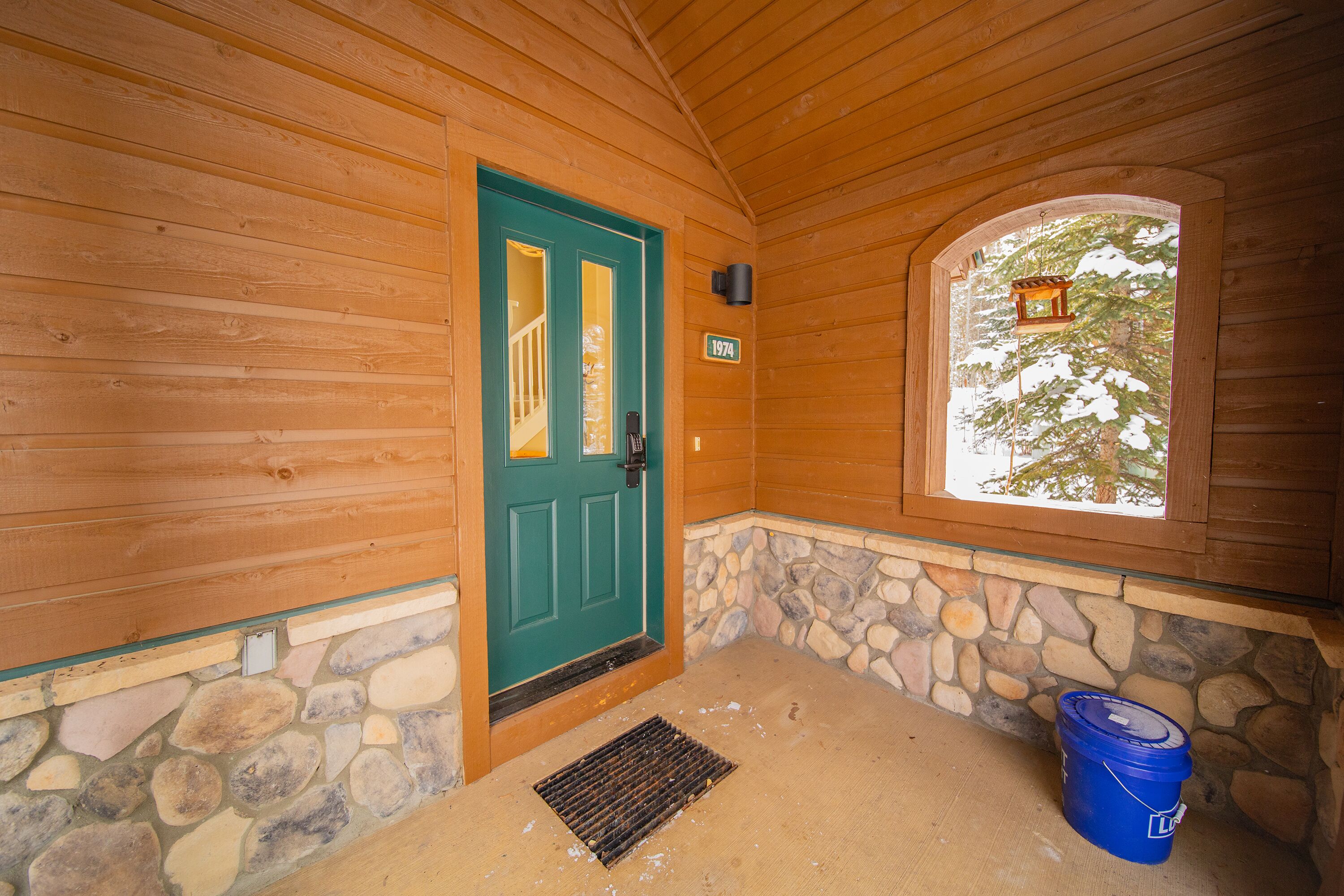 A wooden cabin with a green door, stone foundation, and a blue bucket near the entrance.