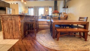 A spacious living area with a wooden ceiling, a central fireplace, a dining table with chairs, and a kitchen area with a refrigerator.