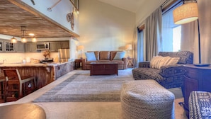 A spacious living area with a large sofa, ottoman, and patterned armchair. A kitchen with wooden cabinets and a bar area is visible.