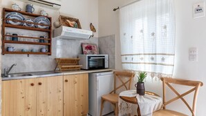 A compact kitchen with wooden cabinets, a microwave, and a small refrigerator. There is a wicker basket on the counter and a chair with a table nearby. - Cottage Cozy Studio (Yianna Cottage Studios) (Skopelos)