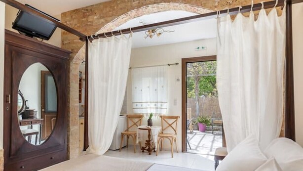 A room with a bed, a TV, a table with chairs, and a view of the outside. - Cottage Cozy Studio (Yianna Cottage Studios) (Skopelos)