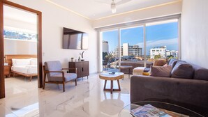 A modern living room with a sofa, armchair, and coffee table, large windows with a city view, and a flat-screen TV mounted on the wall.