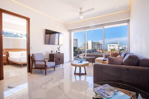 A modern living room with a sofa, armchair, and coffee table, a flat-screen TV mounted on the wall, and large windows offering a city view.