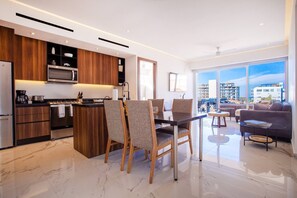 A modern kitchen with a central island, dining chairs, a sofa, and a view of the city through large windows. - Luxe 2BR w/ Balcony, Pool & Gym (Puerto Vallarta)