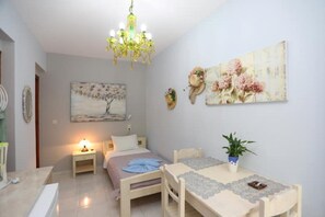Ein kleiner, gut beleuchteter Raum mit einem Bett, einem Nachttisch und einem Tisch mit Stühlen. Eine Kristallleuchter hängt von der Decke, ein Gemälde ziert die Wand und auf dem Tisch steht ein Blumentopf.