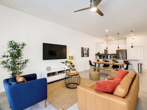A modern living room with a TV, sofa, and coffee table, a dining area with chairs and a table, and a kitchen with appliances. - Fairy Tale Disney Stay / Garage Access / Water Fun (Four Corners)