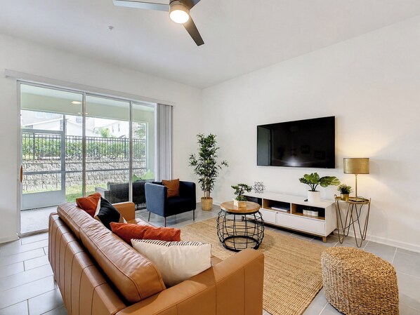 A modern living room with a leather sofa, a TV mounted on the wall, and a large window offering a view of the outdoors.