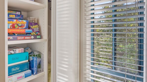 Une pièce avec une fenêtre dotée de stores horizontaux, une bibliothèque remplie de divers jeux de société et une vue sur les arbres à l’extérieur.