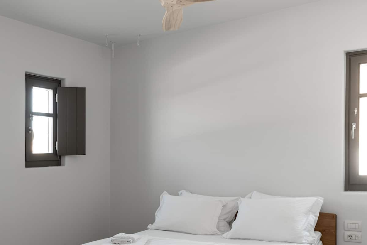 Une chambre moderne avec un cadre de lit en bois, du linge de lit blanc, un ventilateur de plafond et une fenêtre donnant sur un beau paysage.