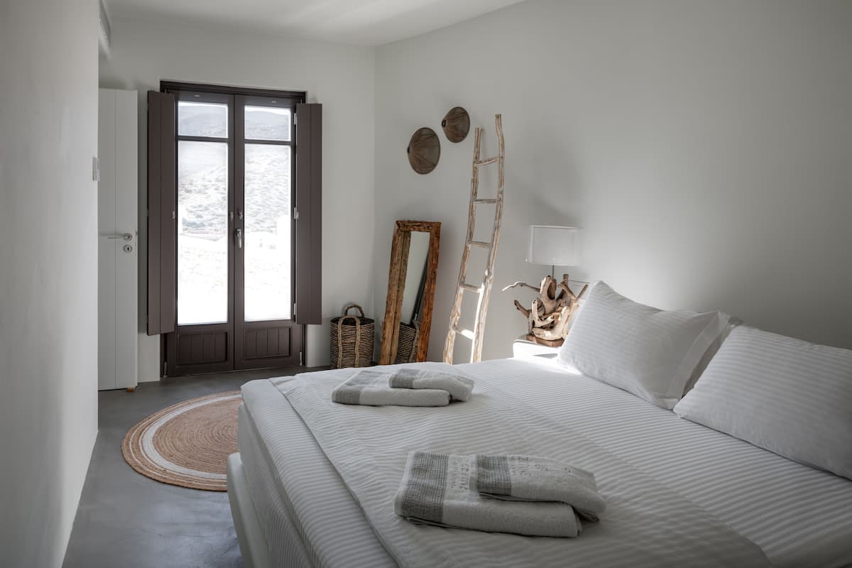 Une chambre à coucher avec un lit, des oreillers, une table de chevet avec une lampe, un miroir, une échelle et une vue sur l’extérieur, visible à travers une porte.