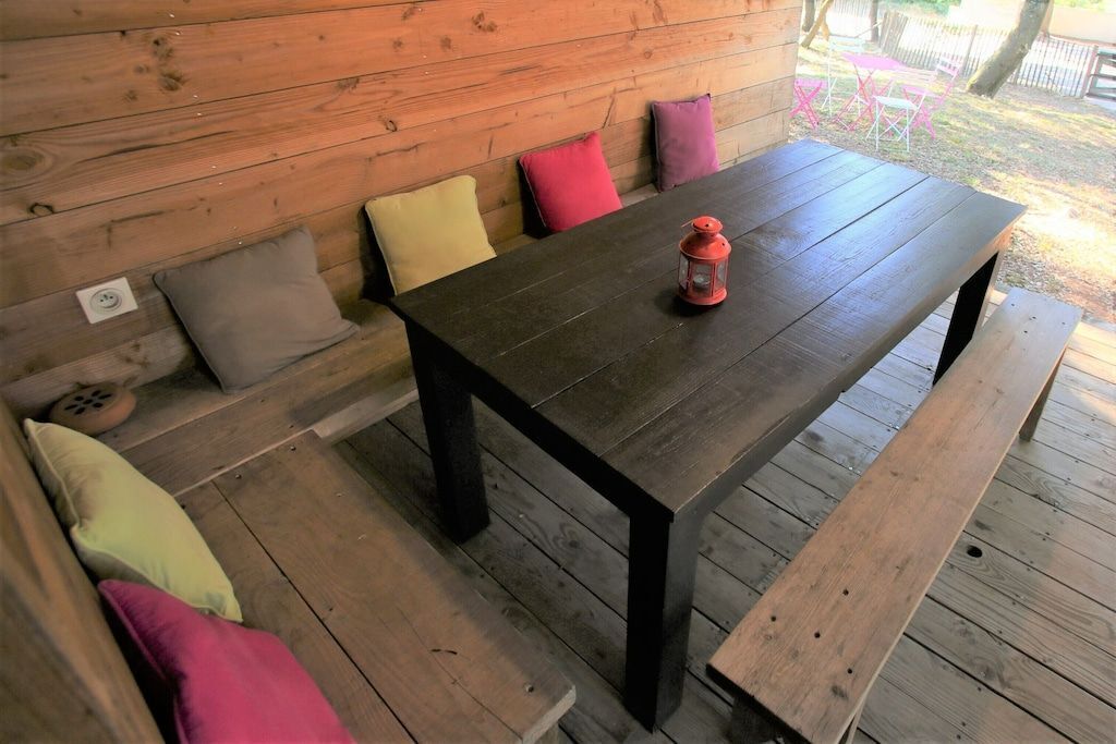 A wooden deck with a dark wooden table and benches, surrounded by cushions.