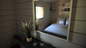 Une petite pièce, de taille modeste, comprenant un lit, un miroir, une fenêtre et une petite table sur laquelle se trouvent une plante et deux verres.
