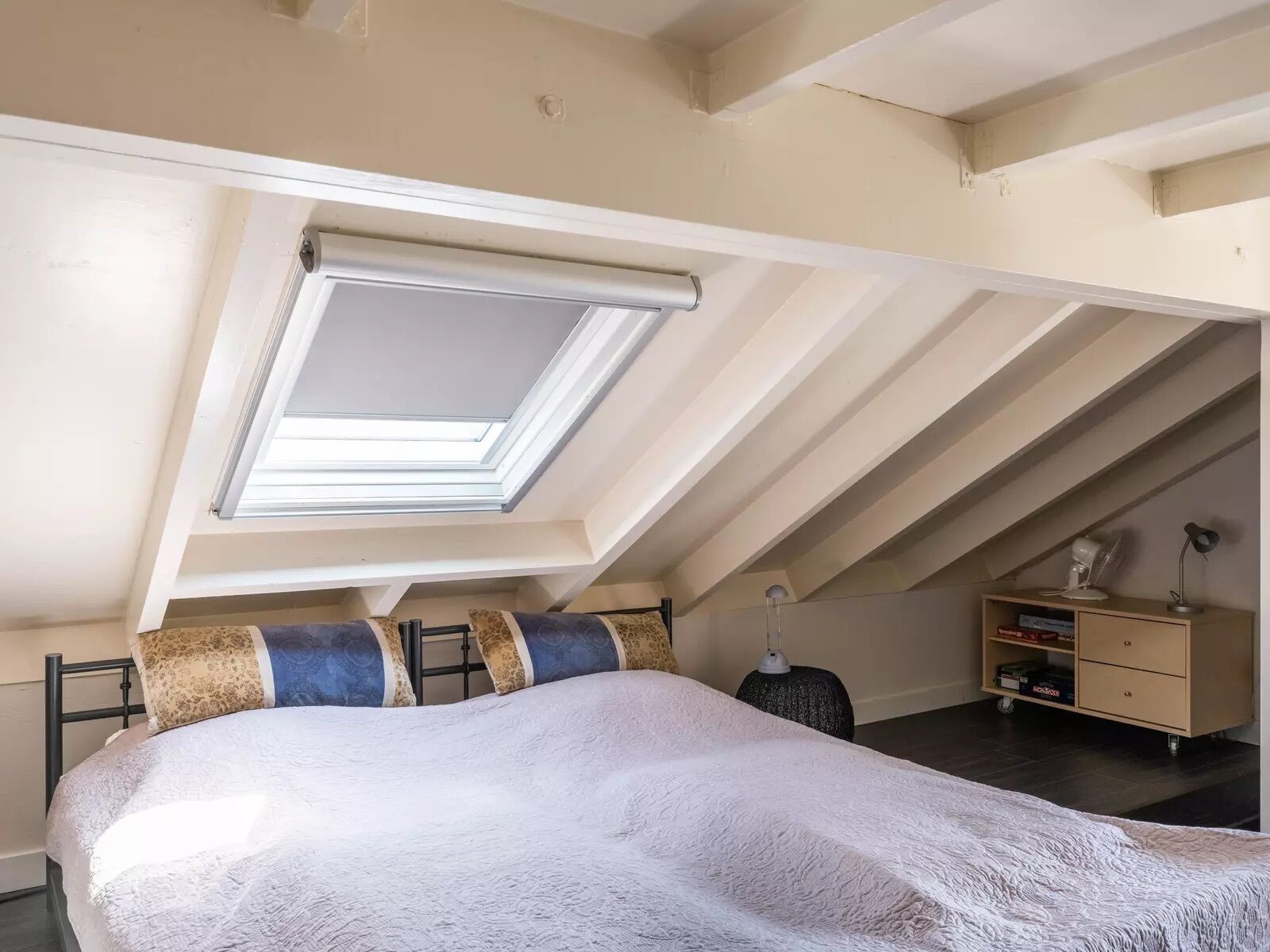 Un dormitorio con tragaluz, una cama con almohadones estampados, una mesita de noche y un pequeño aparador.