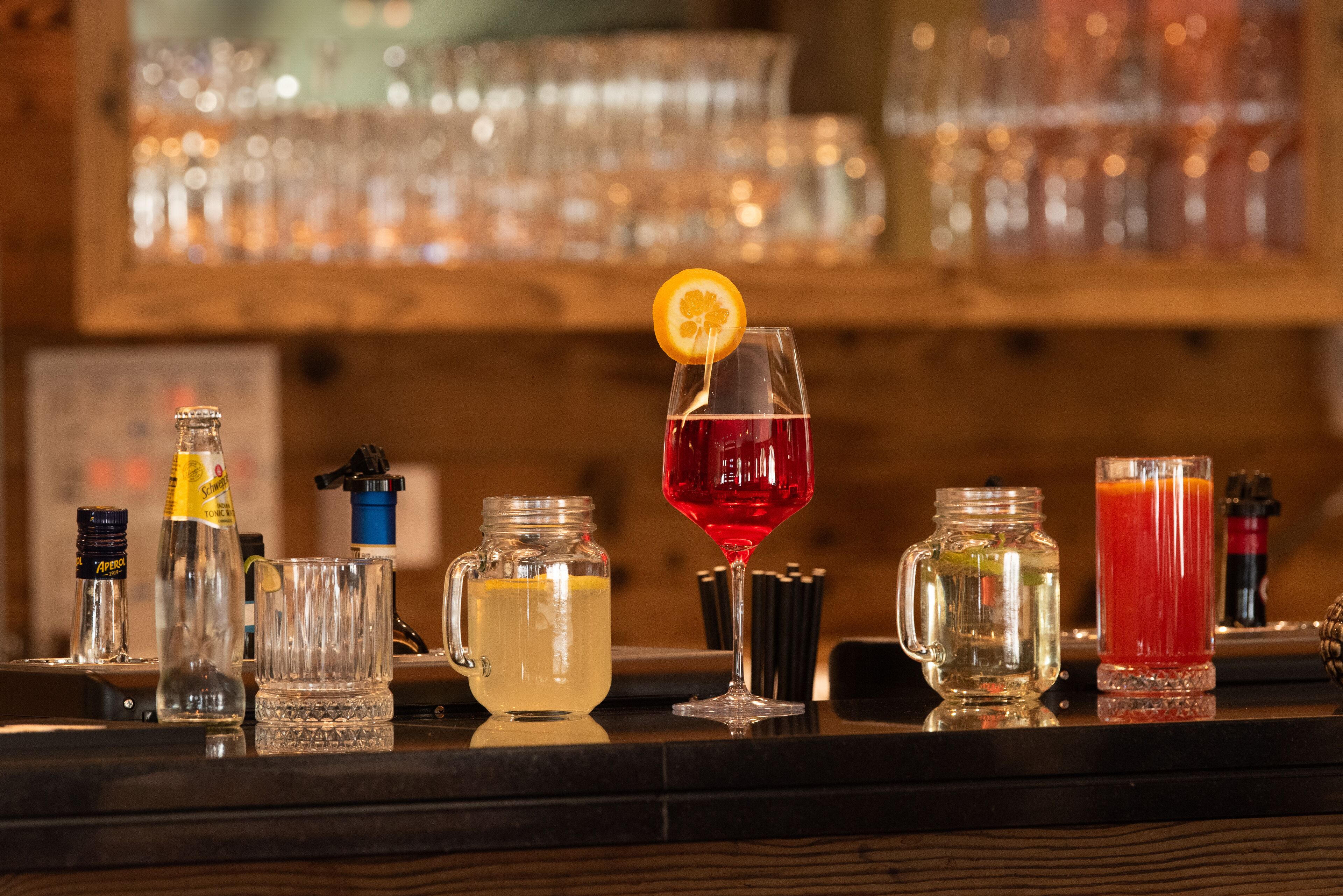 Un bar con diferentes vasos y botellas, incluyendo una bebida roja con un adorno de naranja, un vaso transparente con un trago amarillo y una jarra con una bebida amarilla.