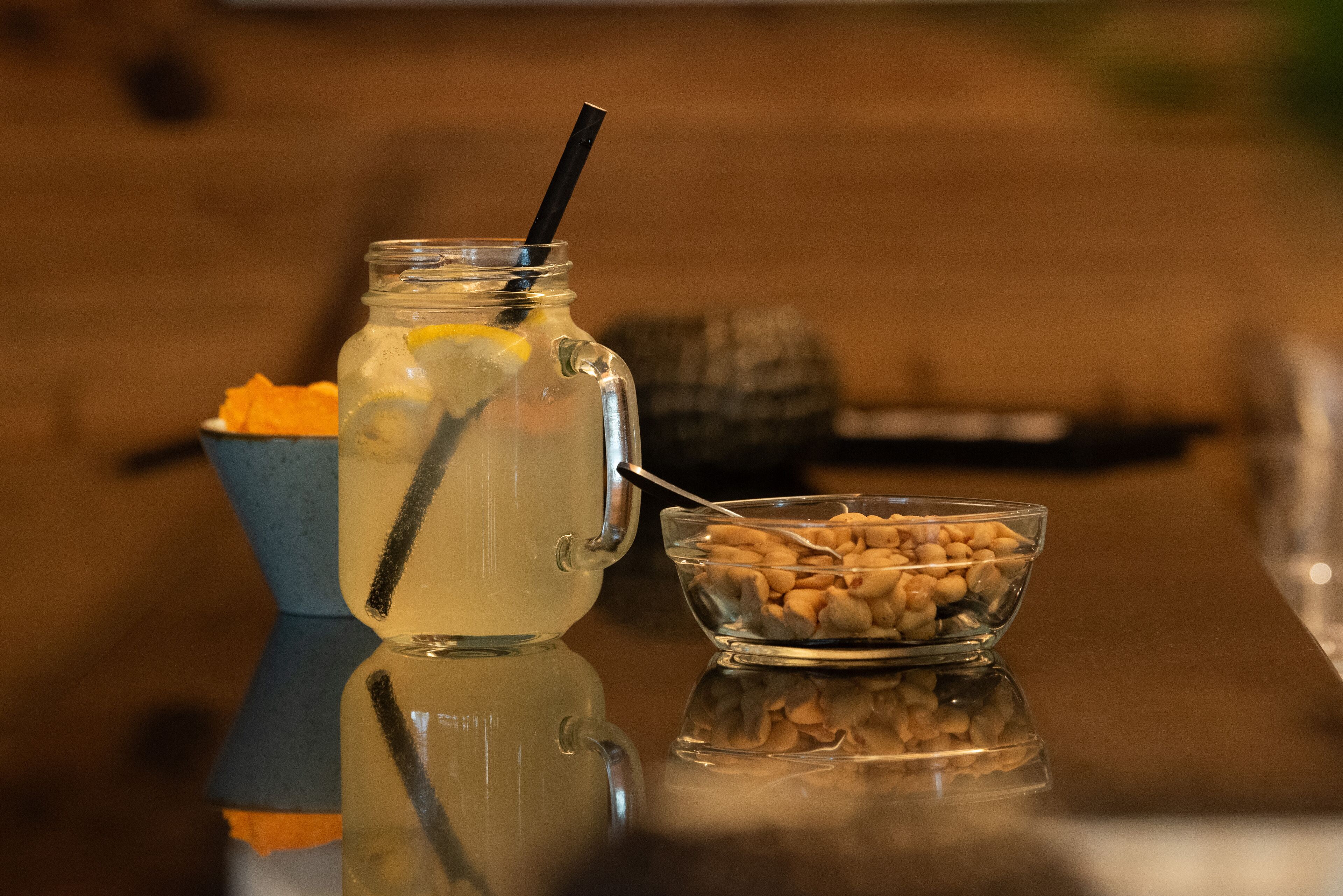A clear glass pitcher with a black straw, a bowl of nuts, and a bowl of orange slices on a reflective surface.