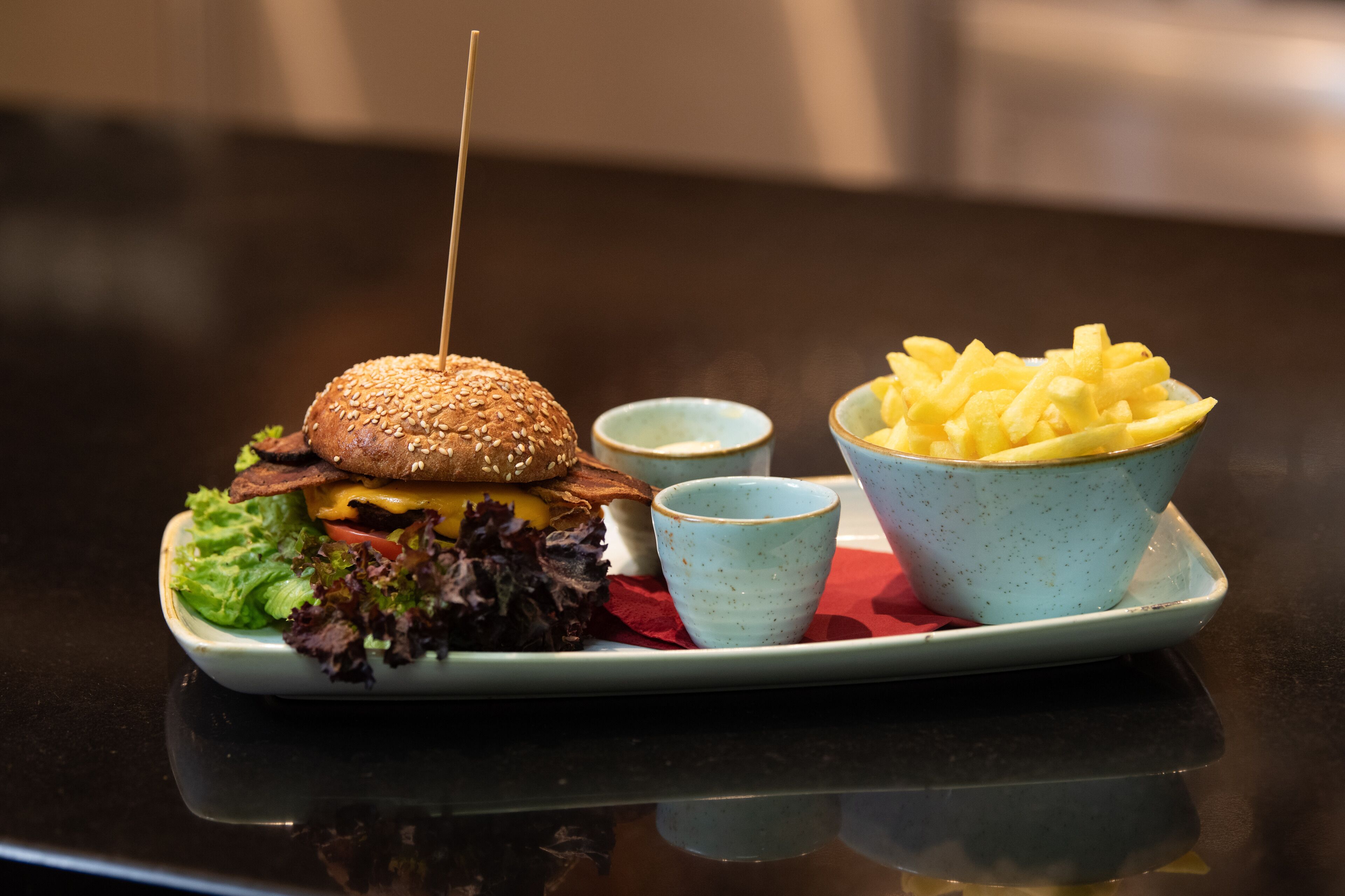 Hamburguesa con palillo, lechuga, tomate, queso y pan con semillas de sésamo, acompañada de papas fritas y dos salsas pequeñas para mojar en un plato.