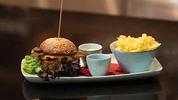 Hamburguesa con palillo, lechuga, tomate, queso y pan con semillas de sésamo, acompañada de papas fritas y dos salsas pequeñas para mojar en un plato.