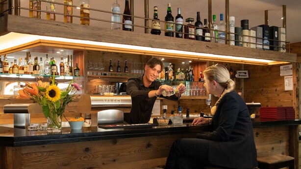 Zona de bar con estantes de madera que exhiben diferentes botellas y copas, una barra y un cliente sentado allí.