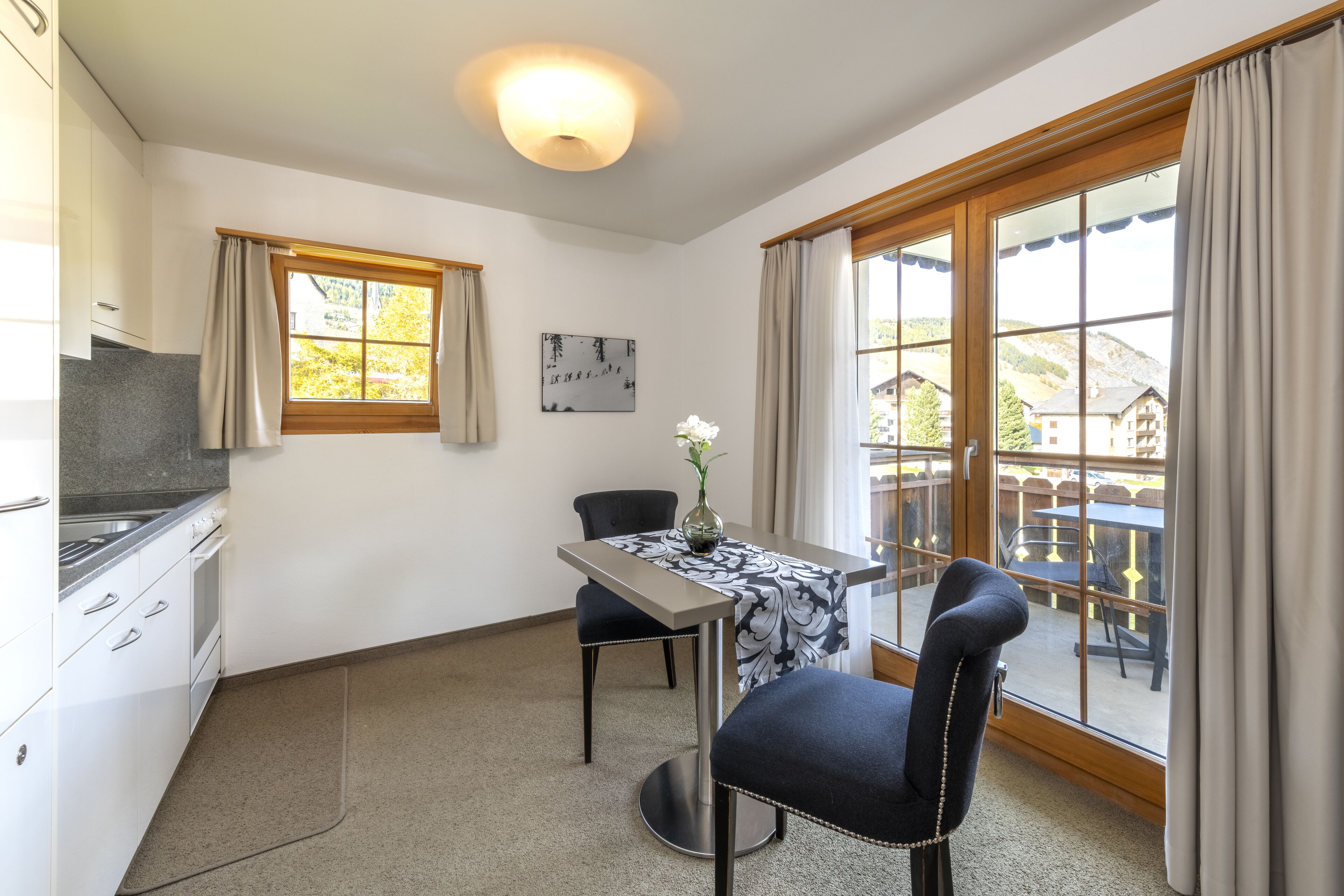 A compact kitchen with white cabinets and a stove, a dining area with a table and chairs, and large windows with curtains.