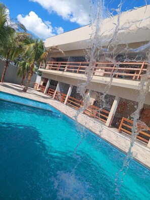 A swimming pool with clear blue water, surrounded by palm trees and a building with balconies. - Alto Costa Flats (Olímpia)