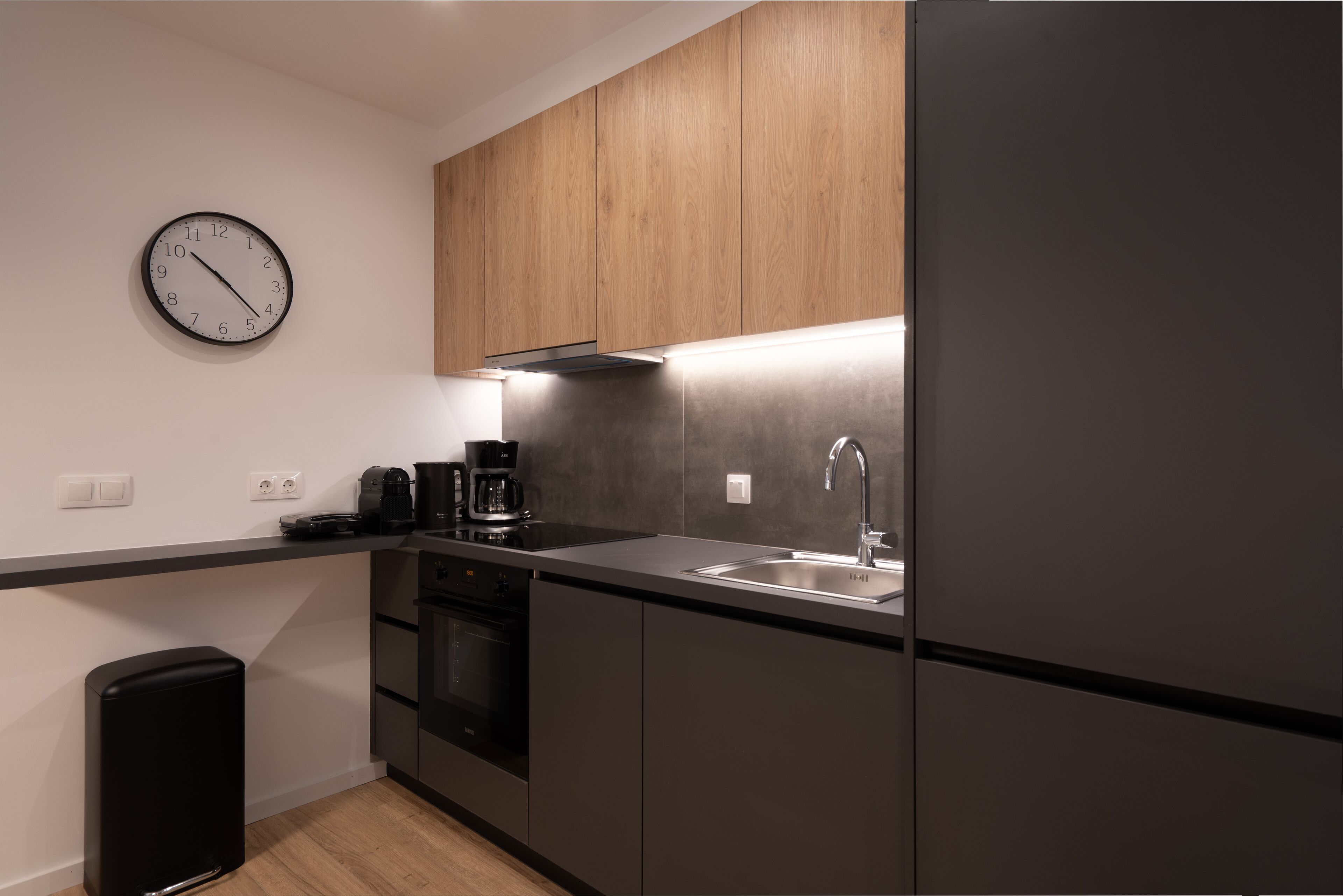 A modern kitchen with dark cabinetry, a stainless steel sink, a coffee maker, and a clock on the wall.