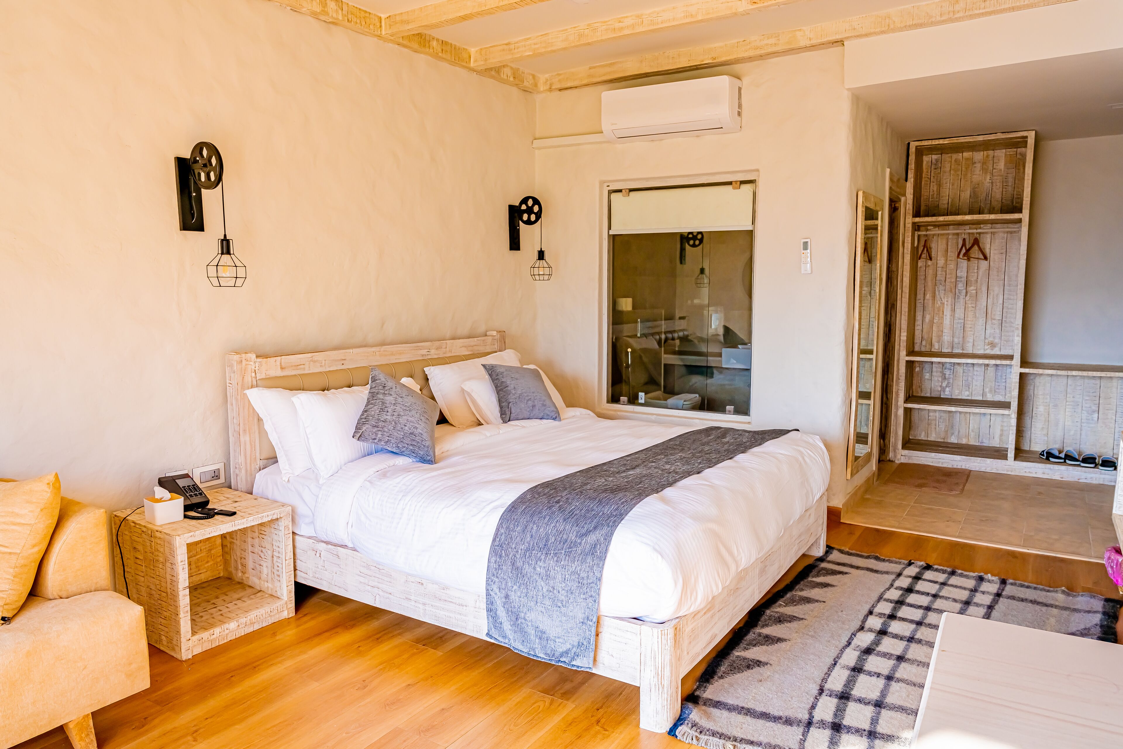 A bedroom with a wooden bed, bedside table, and a small wooden chair. There is a wardrobe with a mirror and a shoe rack.