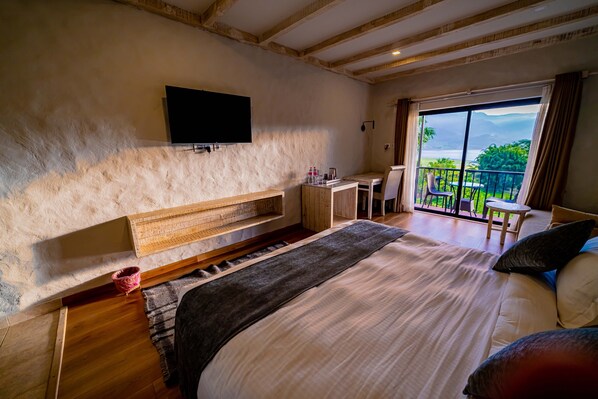 A bedroom with a large bed, a flat-screen TV mounted on the wall, a small desk, and a balcony with a view of the outdoors. - The Hidden Camp (Pokhara)